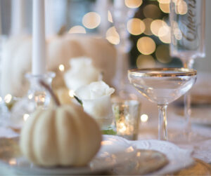 Elegant table setting with white pumpkins, white roses, candles, and bokeh lights in the background. Glassware with gold rims adds a touch of sophistication.