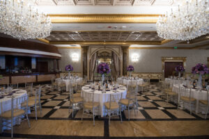A large banquet hall with round tables set for an event, adorned with purple flower centerpieces and surrounded by chairs. Two crystal chandeliers hang from the ceiling, illuminating the room.