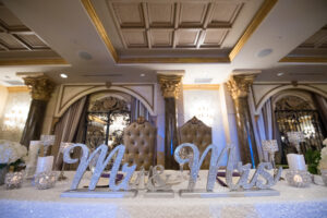 A decorated wedding table with Mr. and Mrs. signs, candles, and light-colored flowers, set in an opulent room with ornate ceiling and pillars. Two high-backed chairs are placed behind the table.