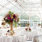 A beautifully decorated, sunlit greenhouse event space that is an ideal site selection for formal gatherings, featuring round tables adorned with vibrant floral centerpieces and white chairs arranged elegantly.