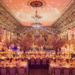 Elegant banquet hall with ornate chandeliers, intricate ceiling designs, and lavishly decorated dining tables adorned with flowers and candles, set for a formal event.