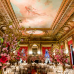 Elegantly decorated banquet hall with ornate ceilings, large floral arrangements, and set dining tables. The ambiance is luxurious with rich red drapery and intricate moldings.