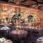 A decorated banquet hall with round tables, tall floral centerpieces, and numerous candles and lights creating an elegant ambiance.