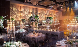 A decorated banquet hall with round tables, tall floral centerpieces, and numerous candles and lights creating an elegant ambiance.