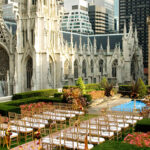 Outdoor wedding ceremony setup on a rooftop garden with chairs arranged in rows, surrounded by greenery and flowers. The background features a historic Gothic-style building and city skyscrapers.