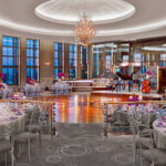 An elegant banquet hall set for an event with round tables, pink floral centerpieces, and a stage with a grand piano and cello. The hall features large windows and a central chandelier.