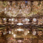 Luxurious banquet hall with ornate chandeliers, floral arrangements, mirrored floor, and elegantly set tables prepared for a formal event.
