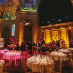 Elegant event space with ornate ceilings and warm lighting, featuring round tables adorned with floral centerpieces and surrounded by chairs. People are visible in the background.