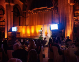 A person stands at a podium on a stage with warm lighting in a large hall, addressing an audience seated at dining tables. Two screens flank the stage, displaying identical content.