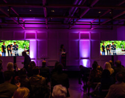 A speaker stands at a podium in a dimly lit room with purple lighting, addressing an audience. Two screens display the text "TRAINING FILMMAKERS" alongside an image of individuals.