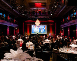 A large gathering of people in a dimly lit, elegant banquet hall featuring a chandelier, with round tables set for dining. A large screen at the front displays a colorful image.