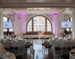 A banquet hall with round tables arranged for an event, featuring white tablecloths, formal place settings, and floral centerpieces. Two large screens display a logo with the word "Justice.