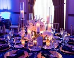 An example of meticulous event planning, this elegant table is adorned with candles, floral centerpieces, glassware, and place settings on a dark tablecloth. The area is dimly lit with blue ambient lighting, creating a sophisticated ambiance.