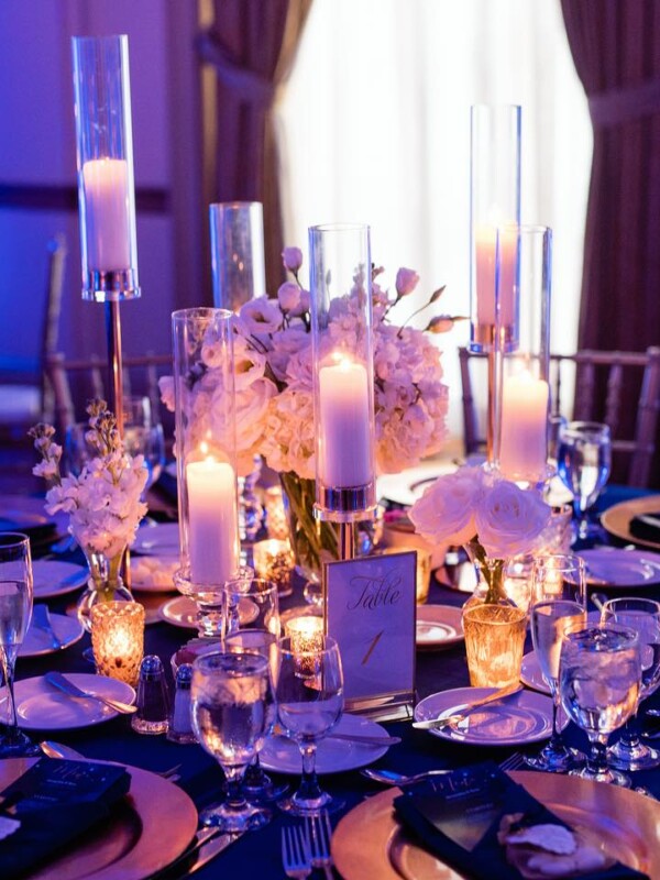 An example of meticulous event planning, this elegant table is adorned with candles, floral centerpieces, glassware, and place settings on a dark tablecloth. The area is dimly lit with blue ambient lighting, creating a sophisticated ambiance.