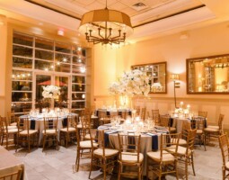 Elegant banquet hall with tables set for an event. The decor includes tall floral centerpieces, candles, and neatly arranged place settings. Warm lighting and large windows create a welcoming ambiance, perfect for seamless event planning.