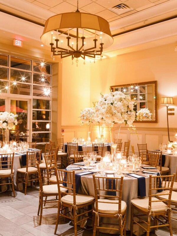 Elegant banquet hall with tables set for an event. The decor includes tall floral centerpieces, candles, and neatly arranged place settings. Warm lighting and large windows create a welcoming ambiance, perfect for seamless event planning.