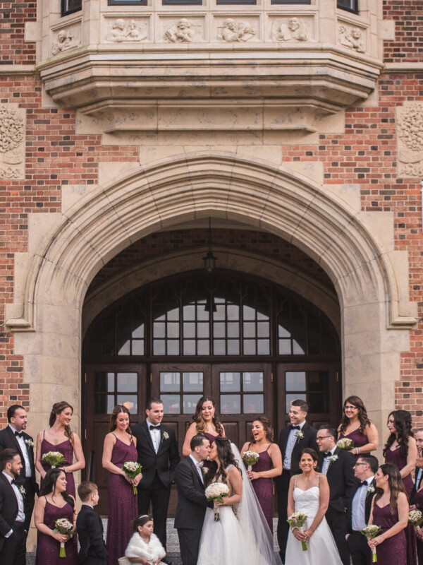 A large wedding party stands in front of an ornate brick building with an arched doorway, perfectly chosen for its charm through meticulous site selection, featuring the bride and groom in the center surrounded by bridesmaids and groomsmen.