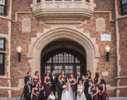 A large wedding party stands in front of an ornate brick building with an arched doorway, perfectly chosen for its charm through meticulous site selection, featuring the bride and groom in the center surrounded by bridesmaids and groomsmen.