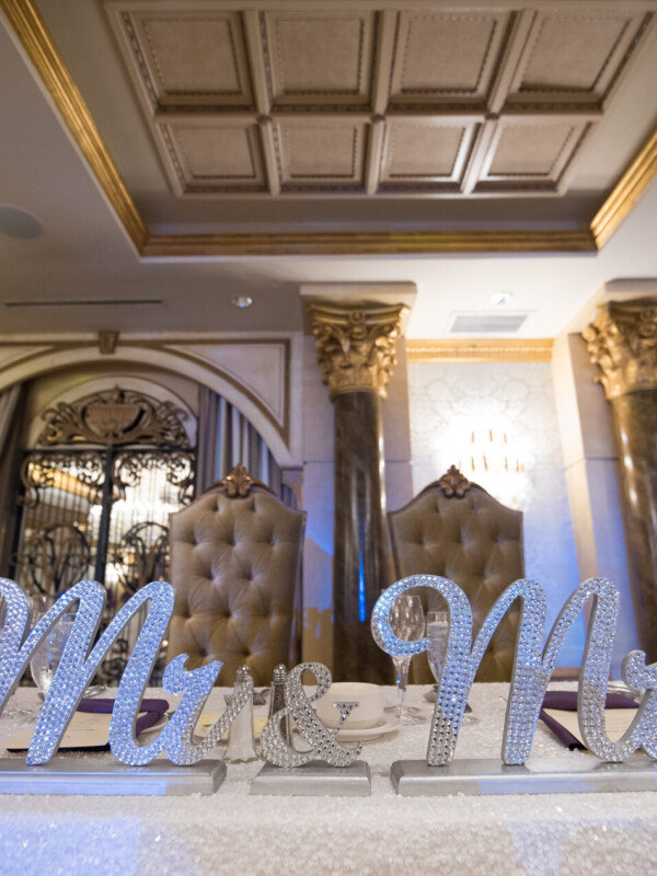 A decorated wedding table with Mr. and Mrs. signs, candles, and light-colored flowers, set in an opulent room with ornate ceiling and pillars. Two high-backed chairs are placed behind the table.