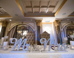 A decorated wedding table with Mr. and Mrs. signs, candles, and light-colored flowers, set in an opulent room with ornate ceiling and pillars. Two high-backed chairs are placed behind the table.