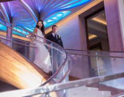 A bride in a white dress and a groom in a dark suit descend a curved staircase with glass railings, under blue and white illuminated ceiling decorations.