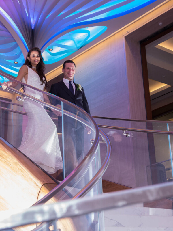 A bride in a white dress and a groom in a dark suit descend a curved staircase with glass railings, under blue and white illuminated ceiling decorations.