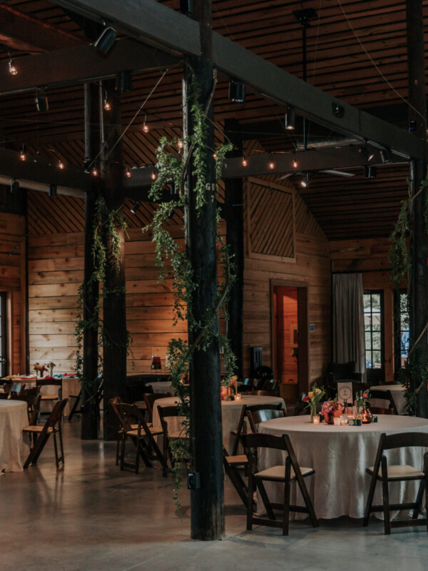 An indoor event space with wooden walls and ceiling, decorated with string lights and greenery. Round tables covered with white tablecloths are set for a gathering.