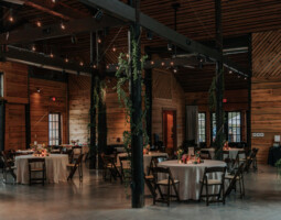 An indoor event space with wooden walls and ceiling, decorated with string lights and greenery. Round tables covered with white tablecloths are set for a gathering.