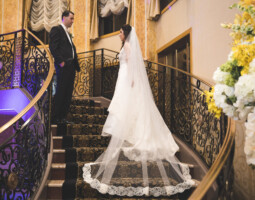 A bride in a white gown and veil stands on a staircase, looking at a groom in a black suit. Ornate railings and a floral arrangement are visible.