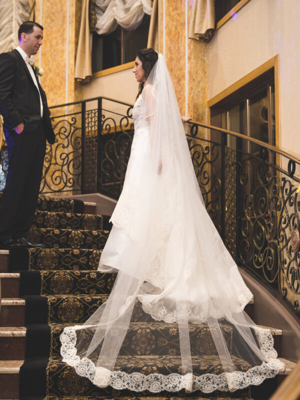 A bride in a white gown and veil stands on a staircase, looking at a groom in a black suit. Ornate railings and a floral arrangement are visible.
