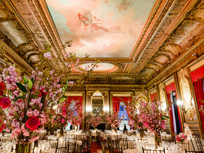 Elegantly decorated banquet hall with ornate ceilings, large floral arrangements, and set dining tables. The ambiance is luxurious with rich red drapery and intricate moldings.