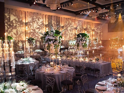 A decorated banquet hall with round tables, tall floral centerpieces, and numerous candles and lights creating an elegant ambiance.