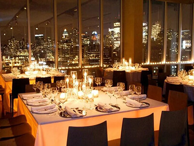 Elegant dining area with white tablecloths and decorative candle lighting, set for an evening event. Large windows offer a view of a city skyline illuminated at night.