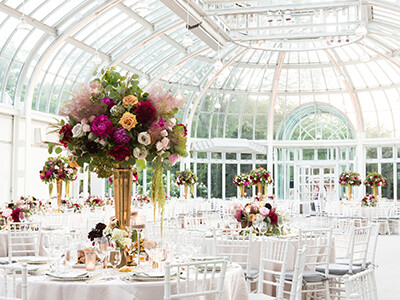 A spacious, elegant event hall with a glass ceiling and large floral centerpieces on round tables decorated for a formal occasion.