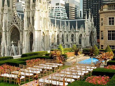 Outdoor wedding ceremony setup on a rooftop garden with chairs arranged in rows, surrounded by greenery and flowers. The background features a historic Gothic-style building and city skyscrapers.