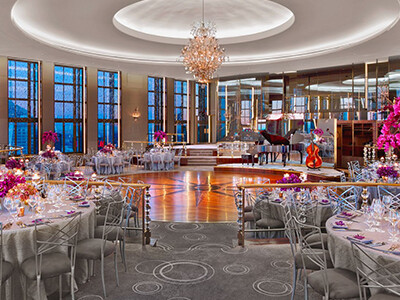 An elegant banquet hall set for an event with round tables, pink floral centerpieces, and a stage with a grand piano and cello. The hall features large windows and a central chandelier.