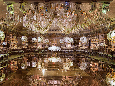 Luxurious banquet hall with ornate chandeliers, floral arrangements, mirrored floor, and elegantly set tables prepared for a formal event.