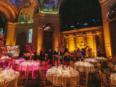 Elegant event space with ornate ceilings and warm lighting, featuring round tables adorned with floral centerpieces and surrounded by chairs. People are visible in the background.